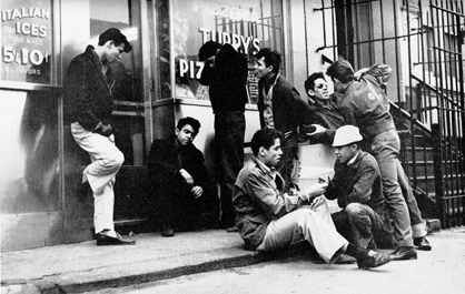 A group of young men hanging outside of a pizza parlor. Some sit on the street while others stand or lean on the wall. Two appear to be pushing one another.